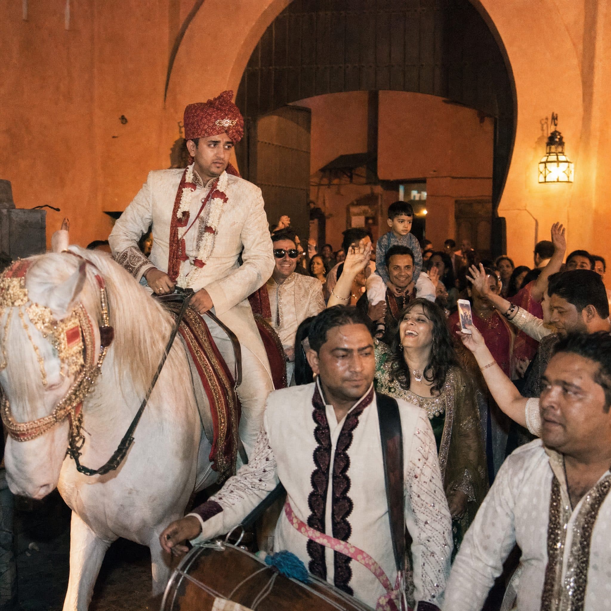Groom arriving by white horse with dhol players and guests during a Baraat at a Marrakech palace gate