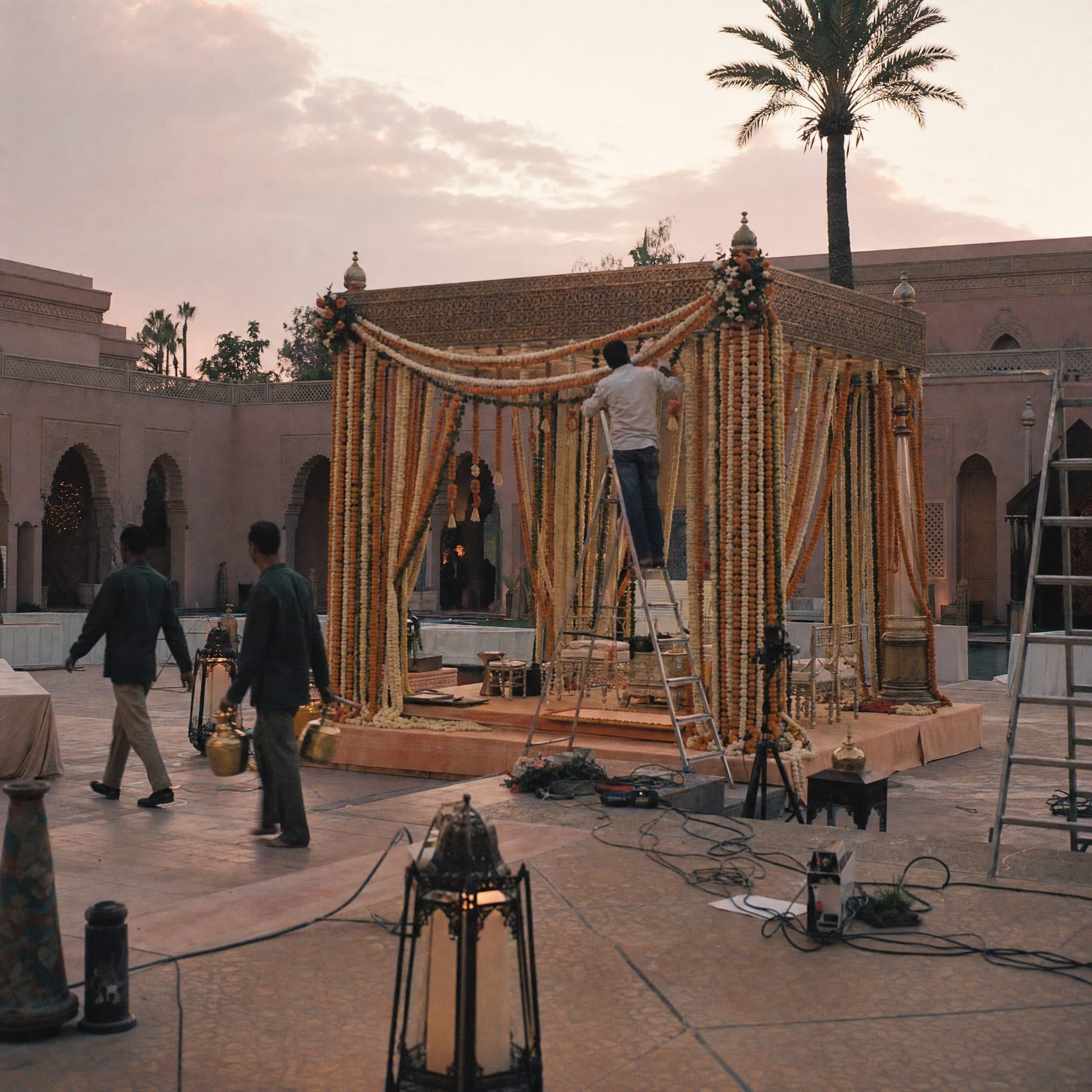 Crew building a marigold-draped mandap in a Marrakech palace courtyard at dawn, with lanterns and cabling still in place